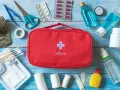 Czerwona apteczka samochodowa z krzyżem i napisem "FIRSTAID" otoczona lekami, bandażami, nożyczkami i innymi akcesoriami medycznymi.