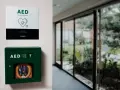 Zielona szafka z defibrylatorem AED na ścianie, obok tabliczka informacyjna.