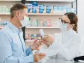 Farmaceutka w masce i okularach podaje pacjentowi w masce pudełko leku.