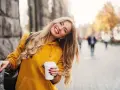 Uśmiechnięta blondynka w żółtej bluzie z kawą w ręku, która przełamuje miejski marazm.