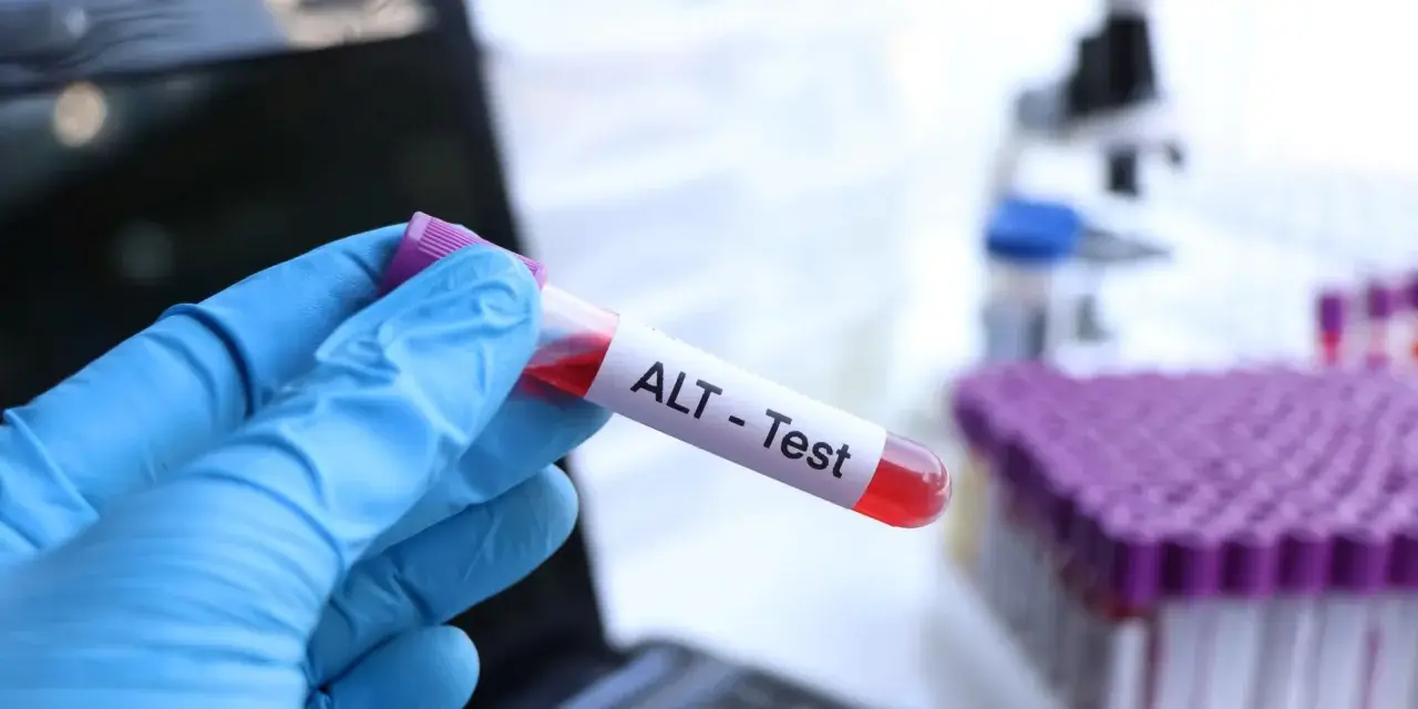 Ręka w niebieskiej rękawiczce trzyma probówkę z krwią oznaczoną "ALT - Test". W tle mikroskop i stojak z probówkami.