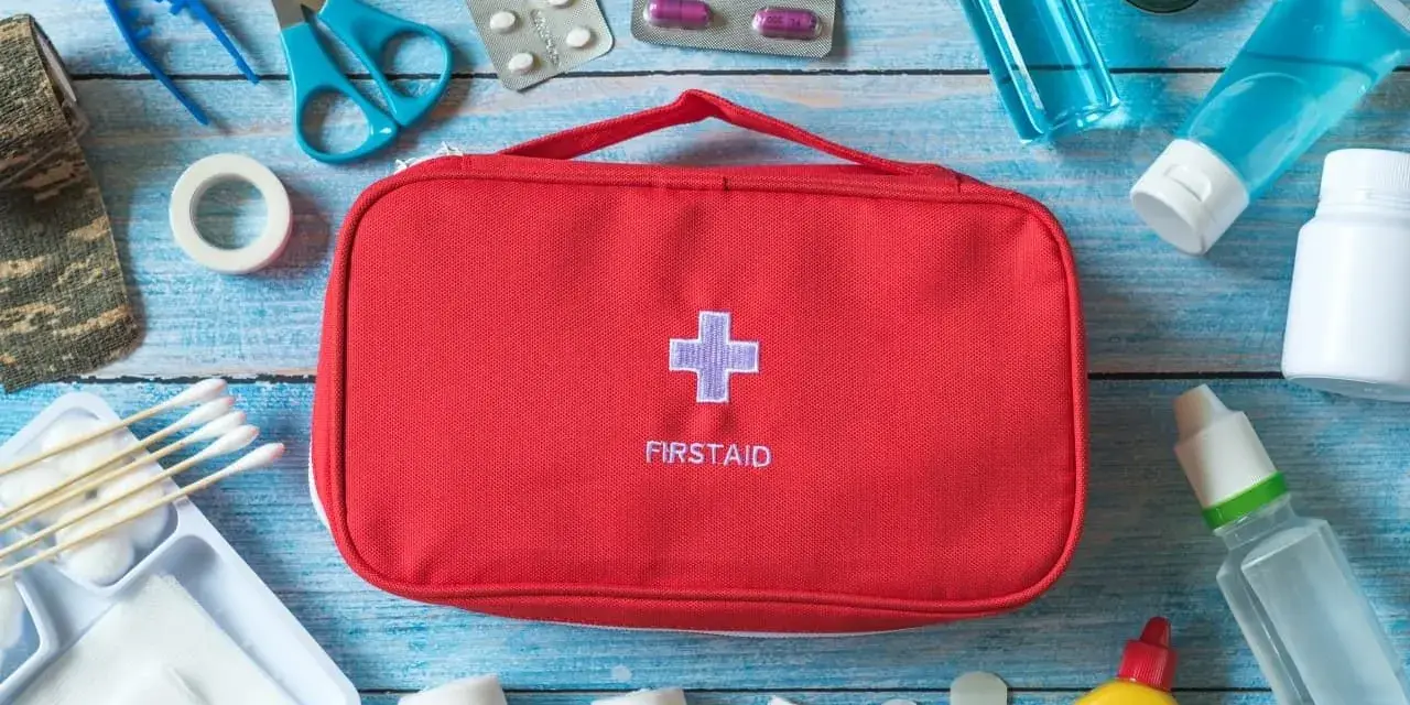 Czerwona apteczka samochodowa z krzyżem i napisem "FIRSTAID" otoczona lekami, bandażami, nożyczkami i innymi akcesoriami medycznymi.