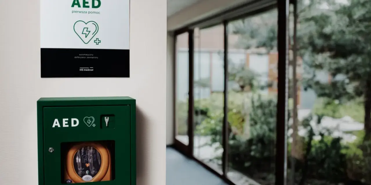 Zielona szafka z defibrylatorem AED na ścianie, obok tabliczka informacyjna.
