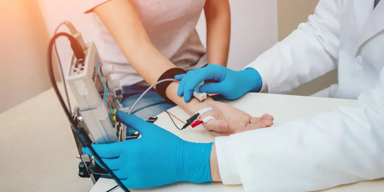Badanie EMG, czyli elektromiografia, to badanie oceniające funkcję nerwów i mięśni. Lekarz z niebieskimi rękawiczkami podłącza elektrody do ręki pacjentki.