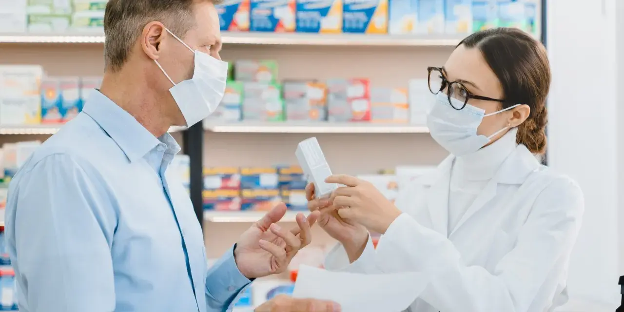 Farmaceutka w masce i okularach podaje pacjentowi w masce pudełko leku.