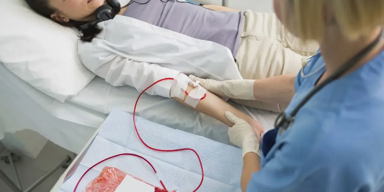 Kobieta leży w łóżku szpitalnym, podłączona do kroplówki z krwią. Pielęgniarka monitoruje zabieg.