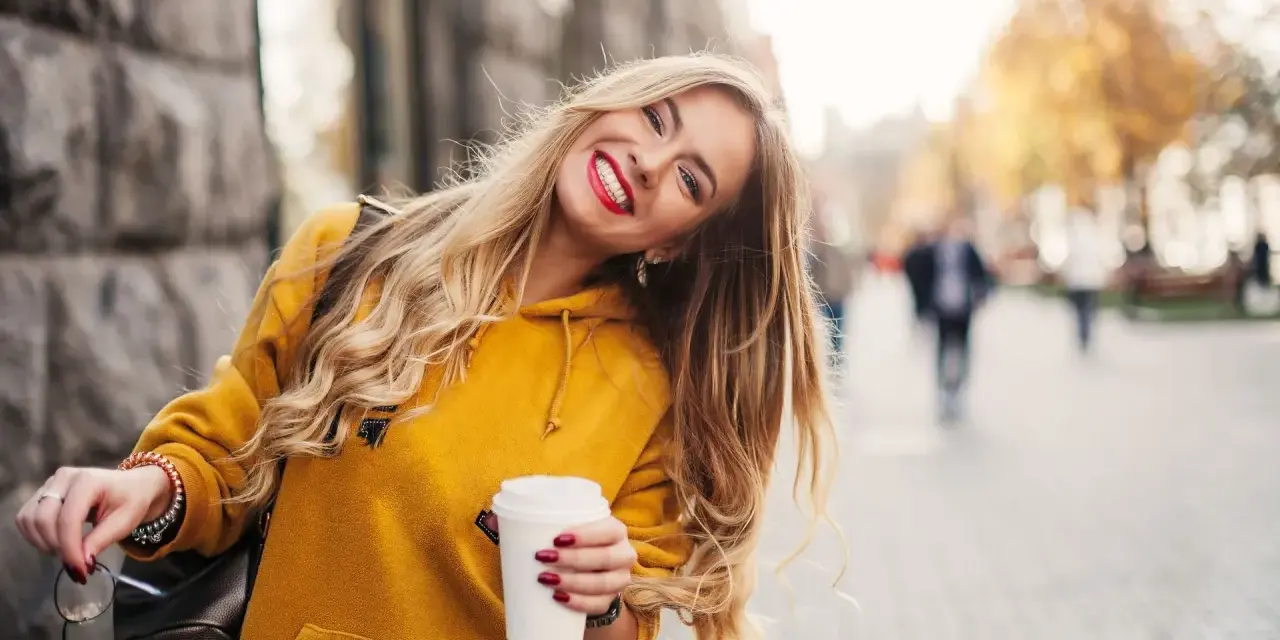 Uśmiechnięta blondynka w żółtej bluzie z kawą w ręku, która przełamuje miejski marazm.