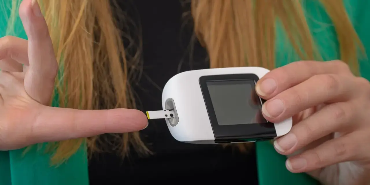Kobieta wykonuje pomiar glukozy glukometrem. Test paskowy jest wsuwany do urządzenia, a palec jest gotowy do pobrania próbki krwi. To ważny element monitorowania krzywej cukrowej.