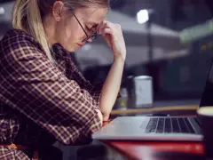Kobieta w okularach z dłońmi na twarzy siedzi przy laptopie, wygląda na zmęczoną.