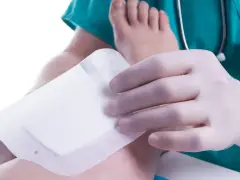 Dłoń w rękawiczce zakłada opatrunek na stopę.