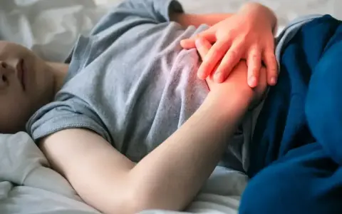 Dziecko leży w łóżku, trzymając się za brzuch. Widać zaczerwienienie sugerujące ból brzucha u dziecka.