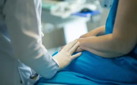 Lekarz w białej rękawiczce kładzie dłoń na ramieniu pacjenta w niebieskiej koszuli.
