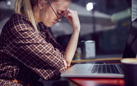 Kobieta w okularach z dłońmi na twarzy siedzi przy laptopie, wygląda na zmęczoną.