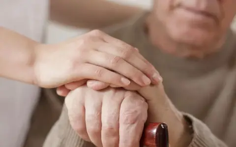 Dłoń opiekunki na dłoni starszego mężczyzny, który trzyma laskę. Wsparcie i troska, jak przy leczeniu np. chorobą Parkinsona, gdzie **Madopar** może pomóc.