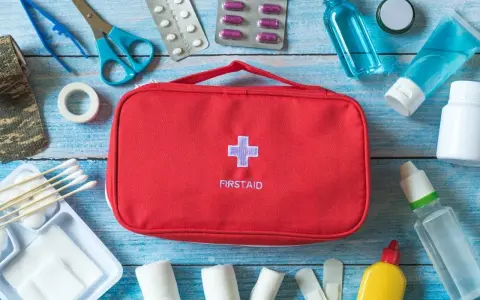 Czerwona apteczka samochodowa z krzyżem i napisem "FIRSTAID" otoczona lekami, bandażami, nożyczkami i innymi akcesoriami medycznymi.