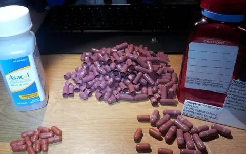 Kapsułki Asacol i inne leki na drewnianym blacie. Wiele różowych kapsułek leży obok butelki Asacol.