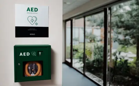 Zielona szafka z defibrylatorem AED na ścianie, obok tabliczka informacyjna.
