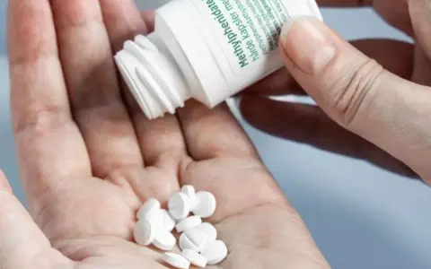 Dłoń rozsypuje białe tabletki z pojemnika. To lek zawierający metylofenidat, znany jako ritalin.