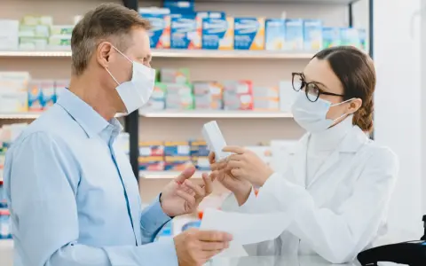 Farmaceutka w masce i okularach podaje pacjentowi w masce pudełko leku.