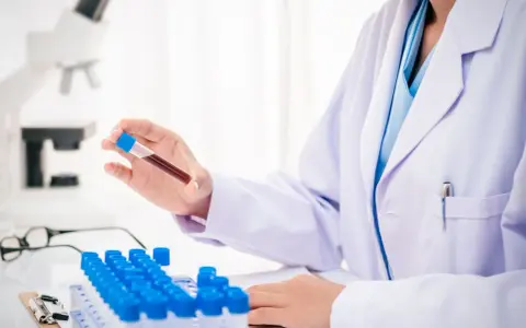 Laborant w białym fartuchu trzyma probówkę z krwią, w tle mikroskop i rząd probówek.