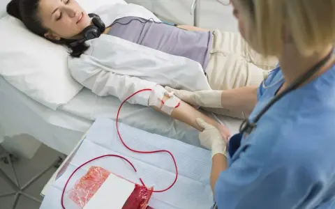Kobieta leży w łóżku szpitalnym, podłączona do kroplówki z krwią. Pielęgniarka monitoruje zabieg.