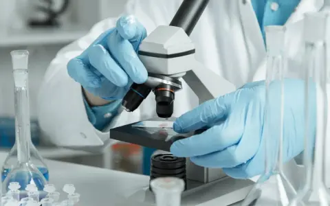 Ręce w niebieskich rękawiczkach badają próbkę pod mikroskopem w laboratorium.