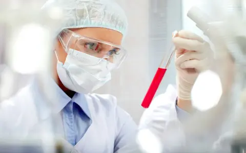 Naukowczyni w okularach i masce bada probówkę z czerwonym płynem. Wyniki wskazują na pct podwyższone.