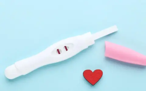 Biały test ciążowy z dwoma czerwonymi paskami, różowa nakładka i czerwone serduszko na niebieskim tle.