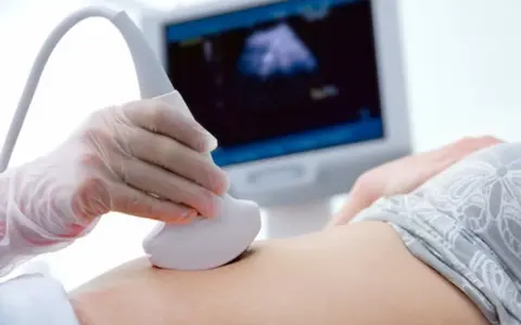 Dłoń w rękawiczce trzyma sondę USG na brzuchu pacjentki, na ekranie widoczny obraz badania.