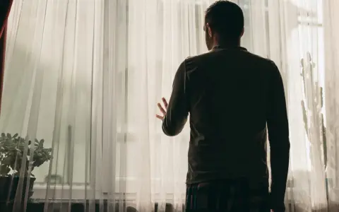 Mężczyzna stoi przy oknie, zasłoniętym przezroczystymi zasłonami. Jego dłoń opiera się o materiał, jakby szukał kontaktu ze światem zewnętrznym, co może być jednym z objawów schizofrenii.