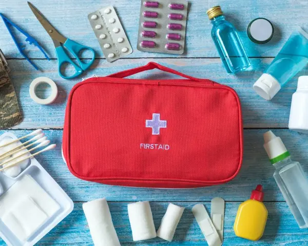 Czerwona apteczka samochodowa z krzyżem i napisem "FIRSTAID" otoczona lekami, bandażami, nożyczkami i innymi akcesoriami medycznymi.