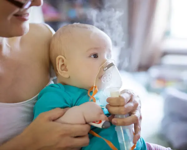 Dziecko z maską inhalatora na twarzy, trzymane przez matkę.