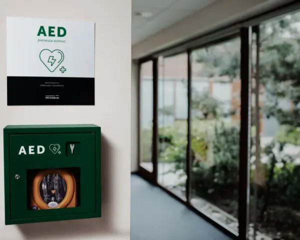 Zielona szafka z defibrylatorem AED na ścianie, obok tabliczka informacyjna.