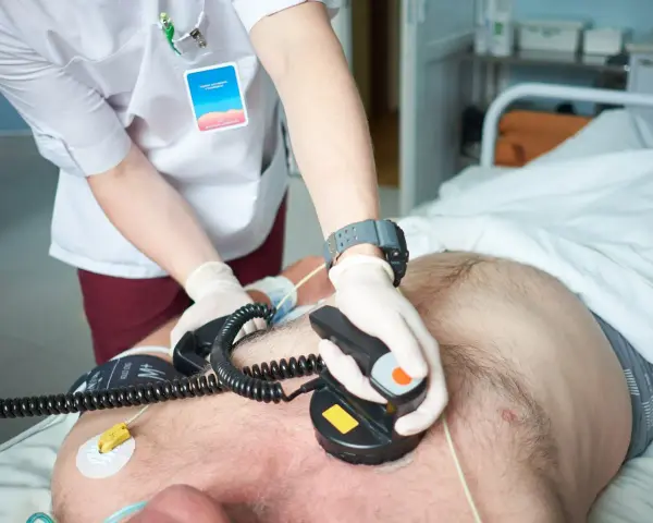 Ratownik medyczny wykonuje kardiowersję pacjentowi leżącemu w łóżku szpitalnym, używając defibrylatora.