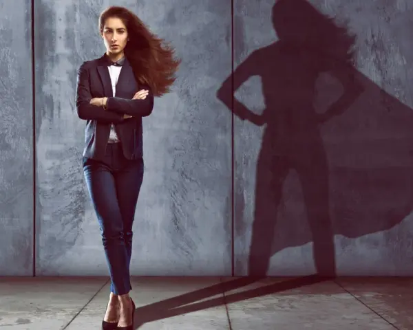 Kobieta w garniturze z rękami skrzyżowanymi na piersi, jej cień na ścianie przypomina superbohaterkę. Czy to oznaka megalomanii?