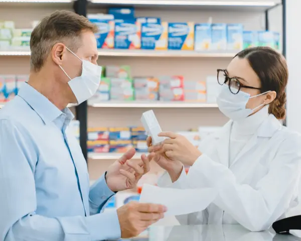 Farmaceutka w masce i okularach podaje pacjentowi w masce pudełko leku.