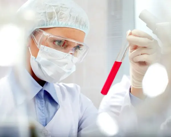 Naukowczyni w okularach i masce bada probówkę z czerwonym płynem. Wyniki wskazują na pct podwyższone.