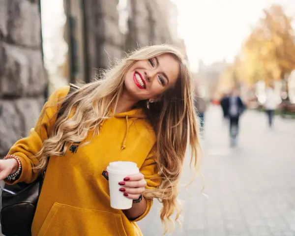 Uśmiechnięta blondynka w żółtej bluzie z kawą w ręku, która przełamuje miejski marazm.