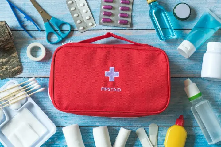 Czerwona apteczka samochodowa z krzyżem i napisem "FIRSTAID" otoczona lekami, bandażami, nożyczkami i innymi akcesoriami medycznymi.