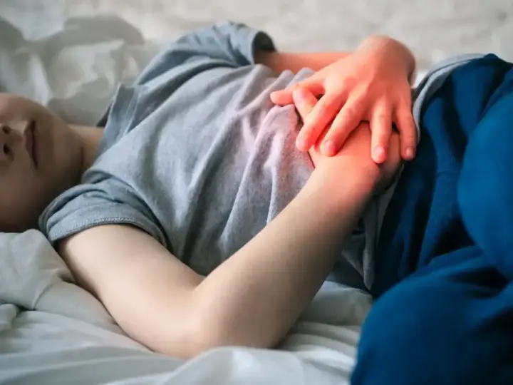 Dziecko leży w łóżku, trzymając się za brzuch. Widać zaczerwienienie sugerujące ból brzucha u dziecka.