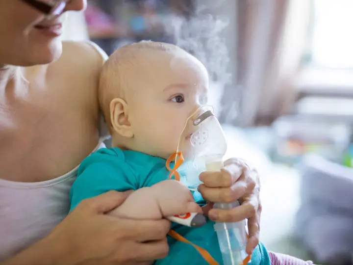 Dziecko z maską inhalatora na twarzy, trzymane przez matkę.