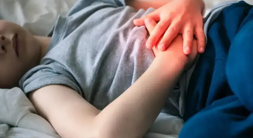 Dziecko leży w łóżku, trzymając się za brzuch. Widać zaczerwienienie sugerujące ból brzucha u dziecka.