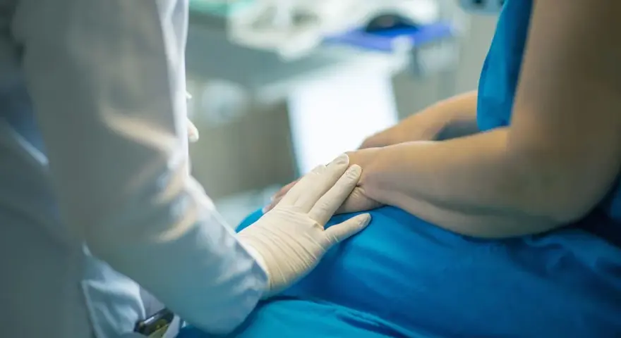 Lekarz w białej rękawiczce kładzie dłoń na ramieniu pacjenta w niebieskiej koszuli.