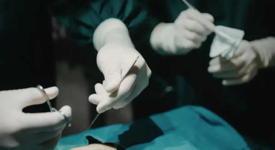 Dłonie chirurga w rękawiczkach trzymające narzędzia chirurgiczne i gazę.