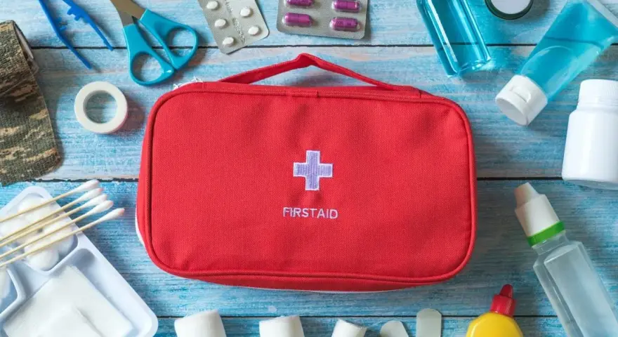 Czerwona apteczka samochodowa z krzyżem i napisem "FIRSTAID" otoczona lekami, bandażami, nożyczkami i innymi akcesoriami medycznymi.