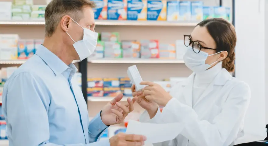 Farmaceutka w masce i okularach podaje pacjentowi w masce pudełko leku.