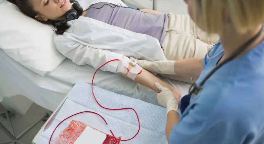 Kobieta leży w łóżku szpitalnym, podłączona do kroplówki z krwią. Pielęgniarka monitoruje zabieg.