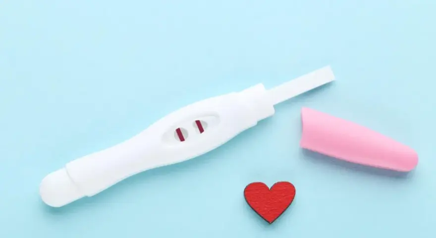 Biały test ciążowy z dwoma czerwonymi paskami, różowa nakładka i czerwone serduszko na niebieskim tle.