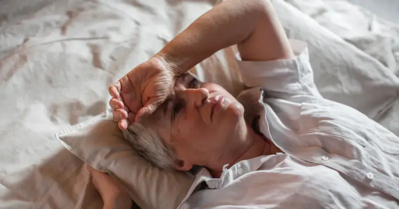 Kobieta leży w łóżku, z ręką na czole, odczuwając uciążliwe perimenopauza objawy.