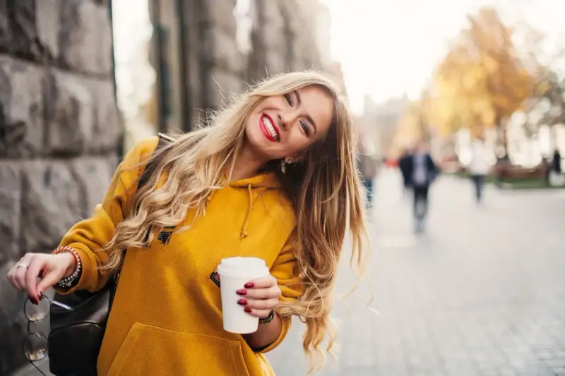 Uśmiechnięta blondynka w żółtej bluzie z kawą w ręku, która przełamuje miejski marazm.